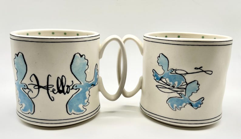 May include: Two white ceramic mugs with a unique handle design, joined together. Each mug features a blue bird design and black text that says "Hello". The rims are decorated with a black line and green dots.