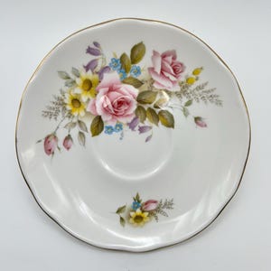 May include: White ceramic saucer with a scalloped edge and gold trim. The saucer is decorated with a floral pattern featuring pink roses, yellow daisies, and blue forget-me-nots. A delicate piece for serving tea or coffee.