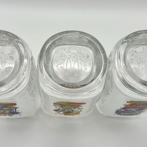 May include: Three clear glass tumblers with a colorful graphic design. The bottom of each glass features the McDonald's logo. The glasses are positioned on a white surface.