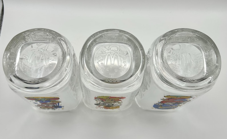 May include: Three clear glass tumblers with a colorful graphic design. The bottom of each glass features the McDonald's logo. The glasses are positioned on a white surface.