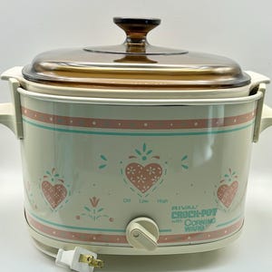 May include: A cream-colored Rival Crock-Pot with Corning Ware insert featuring a pink and teal heart and floral pattern. The appliance has a brown glass lid, side handles, and a front control knob labeled Off, Low, and High with a power cord.
