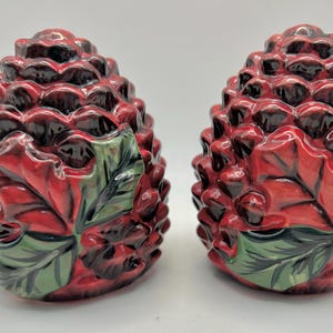 May include: A pair of ceramic salt and pepper shakers shaped like pinecones. The shakers feature a glossy red finish with dark brown shading in the crevices and are decorated with painted green holly leaves and small red berries on the front.