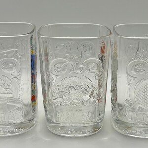 May include: Three clear, square-shaped drinking glasses with embossed designs. The glasses feature various Disney-themed imagery, including stars, fireworks, and the words "Disney Studios". The glasses are empty and sit on a white surface.