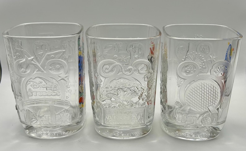 May include: Three clear, square-shaped drinking glasses with embossed designs. The glasses feature various Disney-themed imagery, including stars, fireworks, and the words "Disney Studios". The glasses are empty and sit on a white surface.