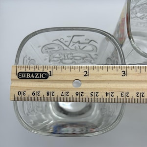 May include: Clear glass square-shaped bowl with etched design, measuring approximately 3 inches across. A wooden ruler is placed across the bowl for measurement. Another glass container is partially visible in the background.