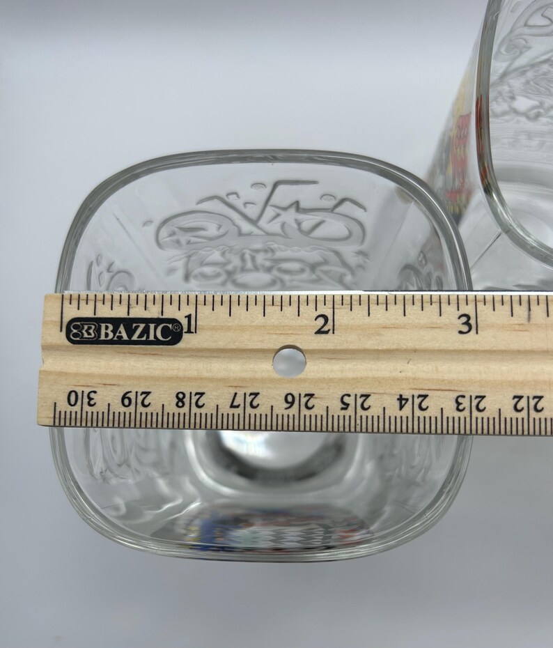 May include: Clear glass square-shaped bowl with etched design, measuring approximately 3 inches across. A wooden ruler is placed across the bowl for measurement. Another glass container is partially visible in the background.