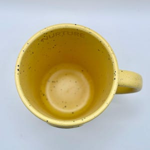 May include: A yellow ceramic mug with a speckled design and a handle. The word "NURTURE" is printed inside the rim of the mug. The mug has a light yellow interior and a slightly darker yellow exterior.