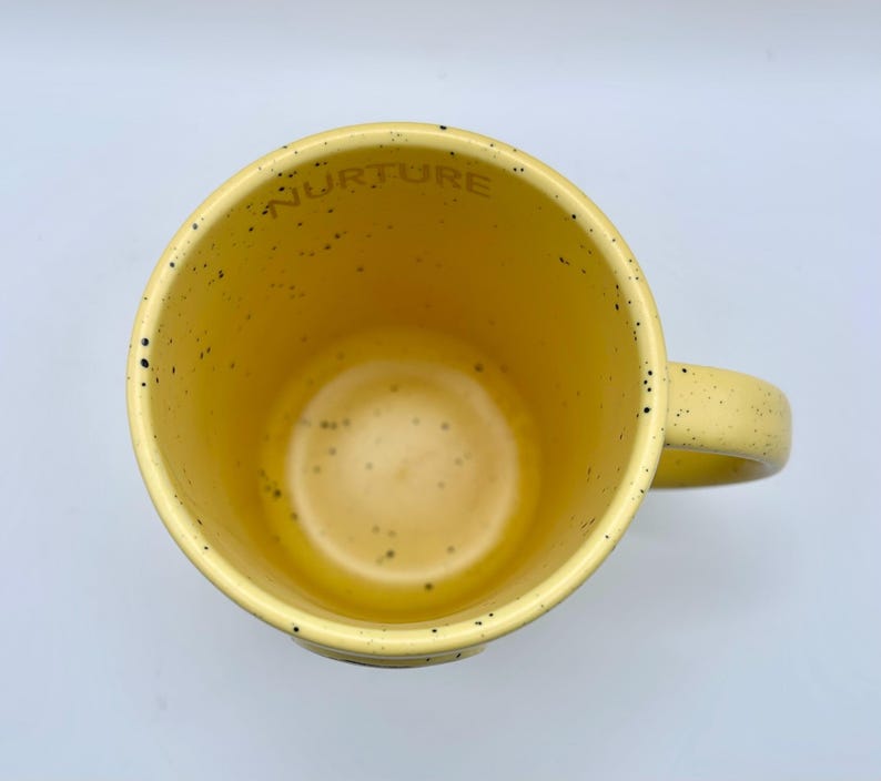 May include: A yellow ceramic mug with a speckled design and a handle. The word "NURTURE" is printed inside the rim of the mug. The mug has a light yellow interior and a slightly darker yellow exterior.