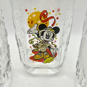 May include: Clear glass tumbler featuring a colorful graphic of Mickey Mouse in safari attire, with a yellow sun and red ribbon design. The word "Disney" is printed below the character, and the number "2000" is at the bottom.