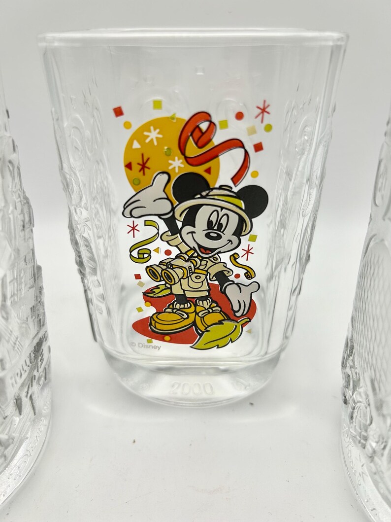 May include: Clear glass tumbler featuring a colorful graphic of Mickey Mouse in safari attire, with a yellow sun and red ribbon design. The word "Disney" is printed below the character, and the number "2000" is at the bottom.