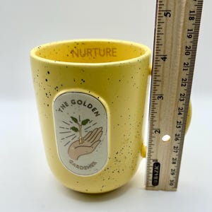 May include: A yellow ceramic mug with black speckles and a handle. The mug features the text "NURTURE" inside the rim and an oval graphic with a hand holding a sprout, with the text "THE GOLDEN GARDENER". The mug is approximately 10 cm tall.