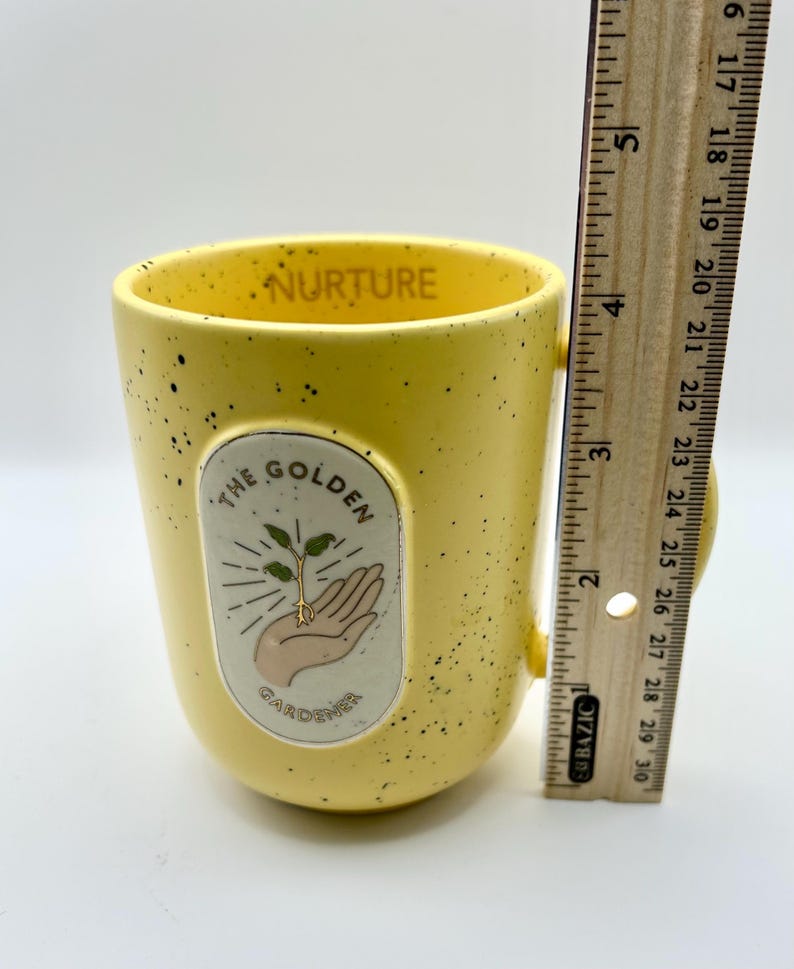 May include: A yellow ceramic mug with black speckles and a handle. The mug features the text "NURTURE" inside the rim and an oval graphic with a hand holding a sprout, with the text "THE GOLDEN GARDENER". The mug is approximately 10 cm tall.
