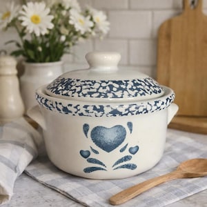 May include: Cream-colored ceramic casserole dish with a lid, featuring a blue heart design and a speckled blue pattern on the rim and lid. The dish has two small handles and is displayed with a wooden spoon and daisies.