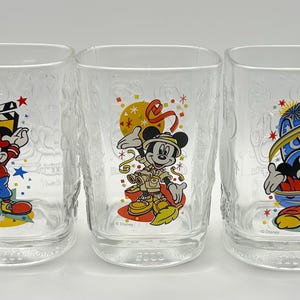 May include: Three clear square-shaped drinking glasses with textured sides. Each glass features a different Mickey Mouse design in vibrant colors. One has a film theme, another a safari theme, and the third a wizard theme. The year 2003 is printed on the bottom.