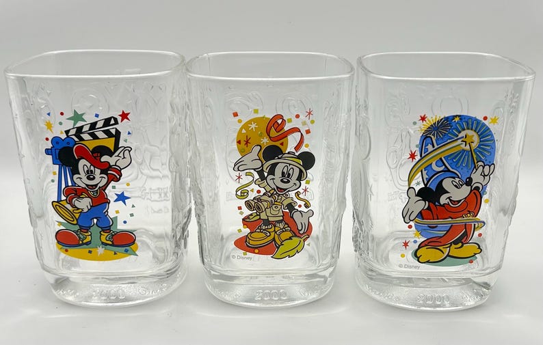 May include: Three clear square-shaped drinking glasses with textured sides. Each glass features a different Mickey Mouse design in vibrant colors. One has a film theme, another a safari theme, and the third a wizard theme. The year 2003 is printed on the bottom.