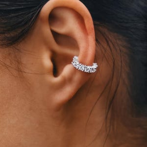 May include: A silver ear cuff adorned with several clear, round gemstones. The cuff is positioned on the ear, highlighting the sparkling stones. This piece of jewellery adds a touch of elegance and is a stylish accessory.
