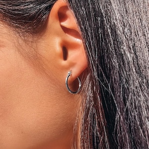 May include: A small, polished silver-toned hoop earring with a circular design and a hinged closure mechanism, worn in the earlobe. The earring features a smooth, reflective metallic surface that catches the light, creating a bright, clean appearance.