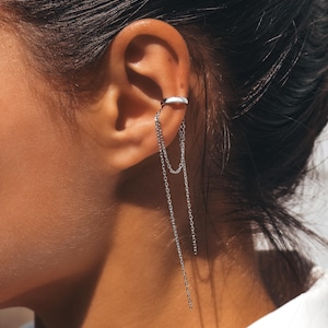 Könnte beinhalten: Silberner Ohrklemme mit baumelnden Ketten. Die Ohrklemme umschließt das obere Ohr und verbindet sich mit zwei zarten Ketten, die herunterhängen. Der Schmuck ist ein schlichtes, elegantes Design.