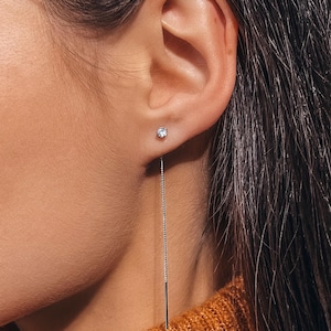May include: A silver-toned threader earring featuring a small, round, clear faceted stone stud at the front, connected to a delicate, thin metal chain that hangs downward, ending in a slender, straight, polished metal bar component.
