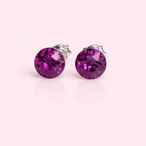 May include: A pair of round, purple stud earrings. Each earring has a faceted, circular gemstone set in a silver-coloured metal backing. The earrings are displayed against a soft pink background.