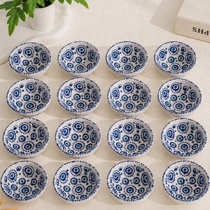 Könnte beinhalten: Ein Set aus sechzehn kleinen, runden Keramikschalen, die in einem vier mal vier Raster angeordnet sind. Jede Schale hat eine weiße Basis mit einem sich wiederholenden blau-dunkelblauen Muster aus konzentrischen Kreisen und Punkten.