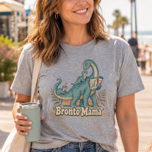 Może przedstawiać: Szary melanżowy t-shirt z grafiką przedstawiającą dużego niebieskiego dinozaura z dwoma mniejszymi dinozaurami, jednym na grzbiecie i drugim na ogonie. Dinozaur trzyma ramkę na zdjęcia i kubek kawy. Poniżej grafiki znajduje się napis "Bronto Mama".