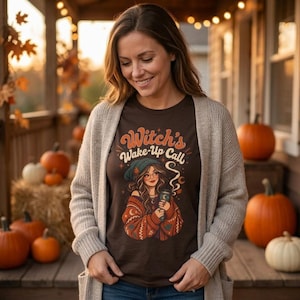 May include: A dark brown t-shirt featuring a graphic illustration of a person wearing a beanie and an oversized jumper while holding a mug. The text "Witch's Wake-Up Call" is printed above the illustration in a stylised, retro-inspired font.