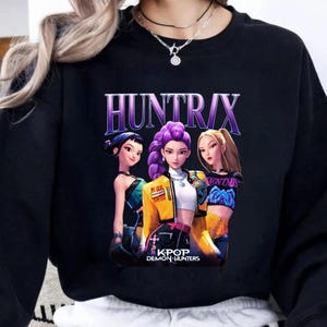 Pode incluir: Camisola preta com um gr&aacute;fico de tr&ecirc;s mulheres animadas com cabelo roxo, loiro e preto. O texto "HUNTRIX" est&aacute; acima das mulheres e "KPOP DEMON HUNTERS" abaixo. As mulheres usam roupas coloridas.