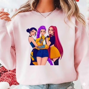 Pode incluir: Um moletom rosa claro com uma ilustra&ccedil;&atilde;o de tr&ecirc;s mulheres. As mulheres, em estilo cartoon, t&ecirc;m cabelo roxo, vermelho e azul escuro e usam roupas estilosas. A figura central usa uma jaqueta amarela e shorts azuis.
