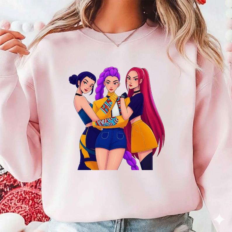 Pode incluir: Um moletom rosa claro com uma ilustra&ccedil;&atilde;o de tr&ecirc;s mulheres. As mulheres, em estilo cartoon, t&ecirc;m cabelo roxo, vermelho e azul escuro e usam roupas estilosas. A figura central usa uma jaqueta amarela e shorts azuis.