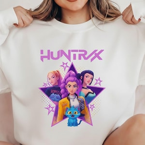Pode incluir: Camisola branca com um gr&aacute;fico de tr&ecirc;s mulheres e um gato azul dentro de uma estrela roxa. A palavra "HUNTRAX" &eacute; impressa acima do gr&aacute;fico em roxo. As mulheres t&ecirc;m cores de cabelo diferentes e usam v&aacute;rias roupas.