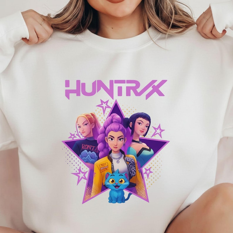 Pode incluir: Camisola branca com um gr&aacute;fico de tr&ecirc;s mulheres e um gato azul dentro de uma estrela roxa. A palavra "HUNTRAX" &eacute; impressa acima do gr&aacute;fico em roxo. As mulheres t&ecirc;m cores de cabelo diferentes e usam v&aacute;rias roupas.