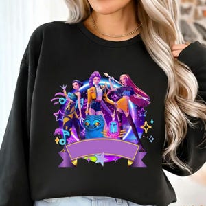 Pode incluir: Camisola preta com um gr&aacute;fico vibrante de tr&ecirc;s figuras femininas estilizadas com um gato azul, num fundo de estrelas de n&eacute;on e notas musicais. Uma faixa roxa e um colar dourado tamb&eacute;m est&atilde;o presentes.