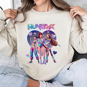 Pode incluir: Camisola creme com um gr&aacute;fico colorido de tr&ecirc;s figuras estilizadas em trajes vibrantes, sobre um fundo em forma de cora&ccedil;&atilde;o. A palavra "HUNTRAX" &eacute; exibida acima das figuras em um gradiente de arco-&iacute;ris.