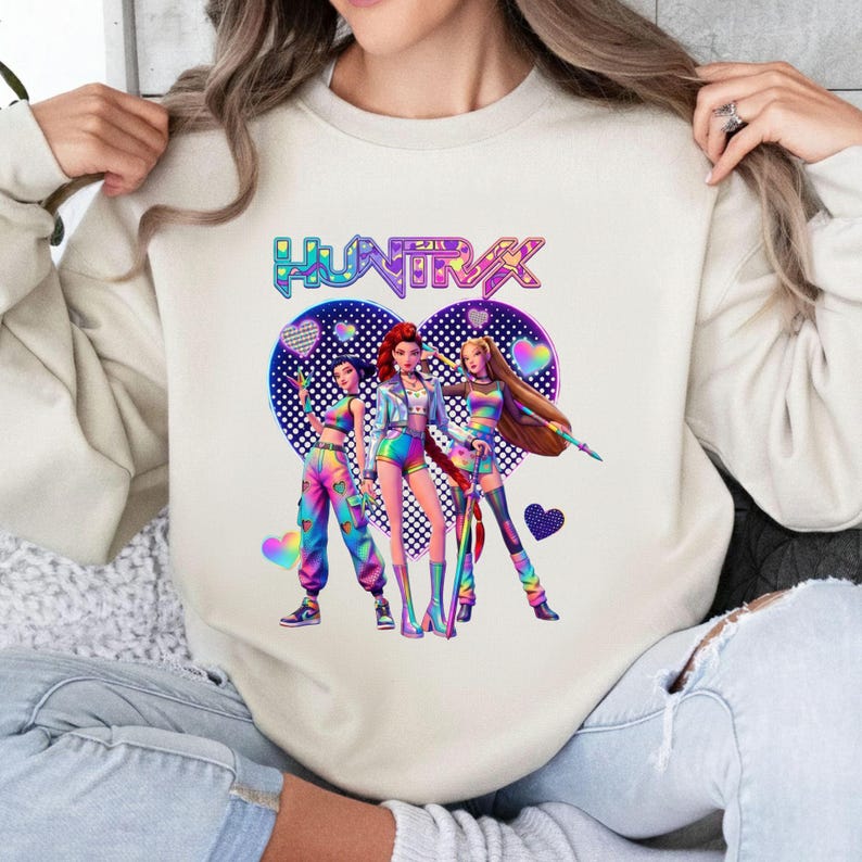 Pode incluir: Camisola creme com um gr&aacute;fico colorido de tr&ecirc;s figuras estilizadas em trajes vibrantes, sobre um fundo em forma de cora&ccedil;&atilde;o. A palavra "HUNTRAX" &eacute; exibida acima das figuras em um gradiente de arco-&iacute;ris.