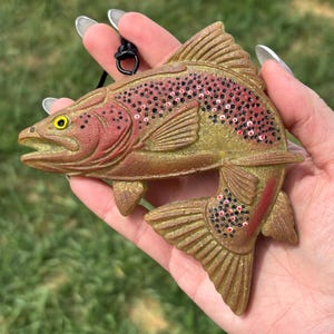 May include: A gold-toned trout ornament featuring a textured body with painted red and black spots along the dorsal area. The fish has a yellow eye, an open mouth, and a black cord loop attached to the top for hanging, showcasing detailed fins.