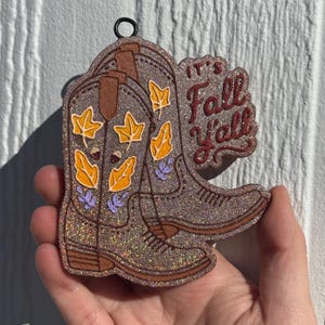 May include: A glittery brown cowboy boot ornament featuring a black hanging loop at the top. The boot is decorated with yellow leaf and small acorn illustrations, along with purple sprigs. The text "IT'S Fall Y'all" is printed in dark red.