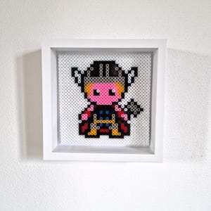 Op de afbeelding: Een ingelijst pixel art-stuk met een cartoonafbeelding van Thor. Het kunstwerk gebruikt zwarte, roze, gele, rode en blauwe kralen om de afbeelding te creëren. De ingelijste kunst is in een witte schaduwbox.