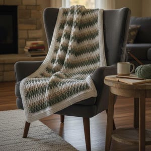 May include: A cozy, hand-crocheted blanket draped over a gray armchair. The blanket features a textured pattern with stripes of white, green, and tan. A white border frames the edges. A mug, yarn, and a book sit on a side table.