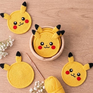 May include: Handmade yellow crochet coasters shaped like Pikachu, featuring black-tipped ears, red cheeks, and smiling faces. Coasters are displayed with a crochet hook and yellow yarn, suggesting a crafting theme.