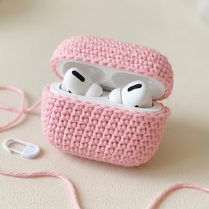 May include: A light pink, crocheted case for wireless earbuds, open to reveal the white earbuds inside. The case is made of a textured yarn and has a matching pink yarn cord and a white safety pin.