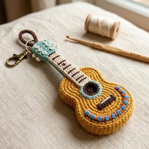 Könnte beinhalten: Gehäkelter Gitarren-Schlüsselanhänger in Gelb-, Blau- und Brauntönen. Das Miniaturinstrument hat ein detailliertes Design mit Schallloch, Saiten und dekorativen Akzenten. Eine braune Schlaufe und ein goldfarbener Verschluss sind angebracht.