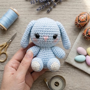 May include: A light blue crocheted bunny with white accents on its feet. The bunny has black eyes, pink cheeks, and a pink nose. The bunny is handmade and is a cute Easter or spring-themed item.