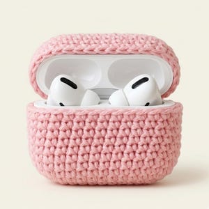 May include: A pink, crocheted case for Apple AirPods Pro. The case is open, revealing white AirPods inside. The case is made of a soft, textured yarn, and the lid is slightly ajar. The background is a solid, neutral color.