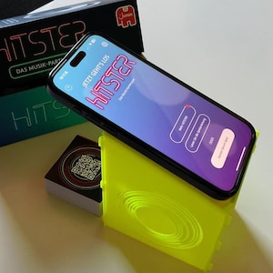 Könnte beinhalten: Ein Smartphone mit der HITSTER-App liegt auf einem leuchtend gelben Ständer. Die App-Oberfläche zeigt das Wort "HITSTER" in pinkfarbenen Neonbuchstaben. Eine Box mit dem Spiel-Logo und ein Kartenspiel sind ebenfalls zu sehen.