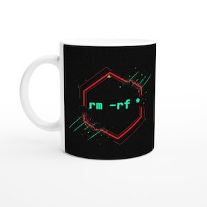 May include: White ceramic mug with a black design featuring the text "rm -rf *" inside a red hexagonal frame. The design includes green and teal accents, and a small yellow triangle. The mug has a white handle.