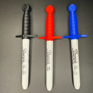 May include: Three Sharpie Fine Point permanent markers with sword-shaped handles in black, red, and blue. The white barrels of the markers display the Sharpie logo and text. The handles are designed to resemble miniature swords.