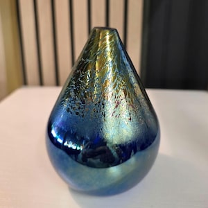 May include: A teardrop-shaped glass vase with a blue and gold iridescent finish. The vase has a narrow opening and a smooth, reflective surface. The color transitions from blue to gold near the top.