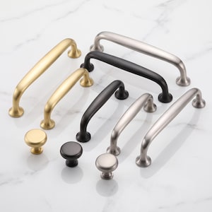 Può includere: Una collezione di ferramenta per mobili in metallo che include diverse maniglie ad arco per cassetti e pomelli rotondi con finiture in oro spazzolato, nero opaco e argento spazzolato. I pezzi sono disposti in gruppo, mostrando varie lunghezze.