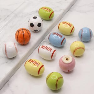 May include: A collection of eleven ceramic cabinet knobs shaped like various sports balls, including basketballs, soccer balls, baseballs, and footballs in colors like orange, white, yellow, blue, pink, and lime green, with one knob showing a metal screw hole.